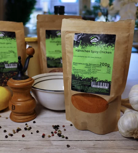 Vier Gewürztüten von Pikant Deutschland auf rustikalem Holzuntergrund, im Vordergrund ‚Hähnchen Spicy Chicken‘ (200g) mit einer detaillierten Zutatenliste: rote Paprika, Salz, Chili, Zucker und Knoblauch. Daneben ein Holz-Pfeffermühle, eine weiße Schale mit schwarzem Rand, Knoblauchknollen, eine Zitrone und verstreute Pfefferkörner. Der Hintergrund zeigt weitere Gewürzverpackungen mit grünen Etiketten.